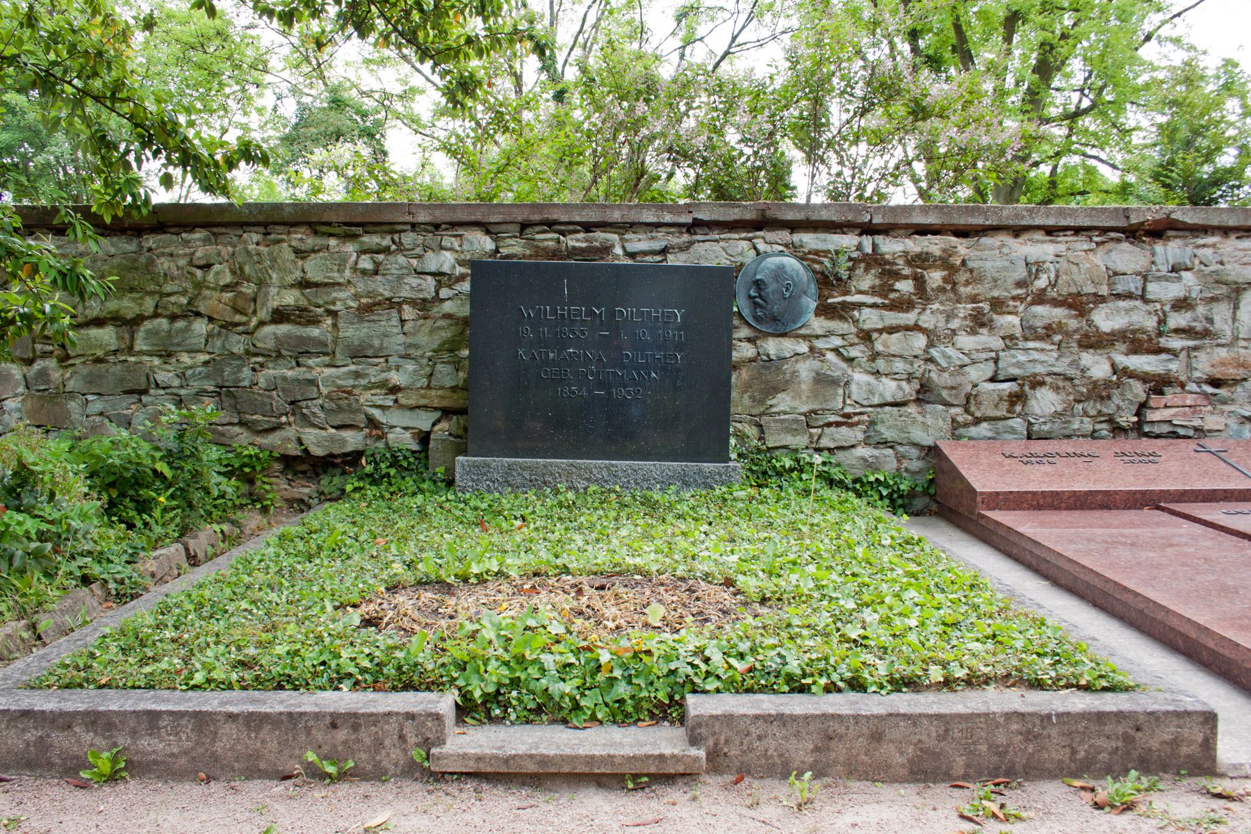 Grab von Wilhelm und Katharina Dilthey mit schwarzer Namenstafel und Porträtrelief vor einer Bruchsteinmauer auf Friedhof Biebrich