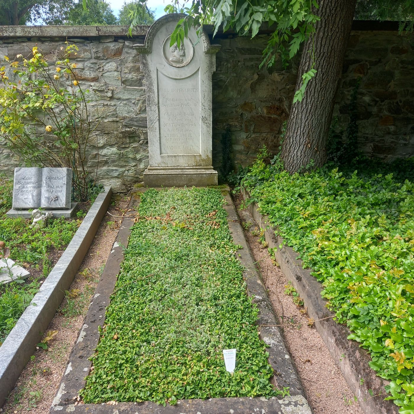 Historische Grabstele Emil Hopfgartens mit rundem Reliefmedaillon vor der alten Bruchsteinmauer auf Friedhof Biebrich