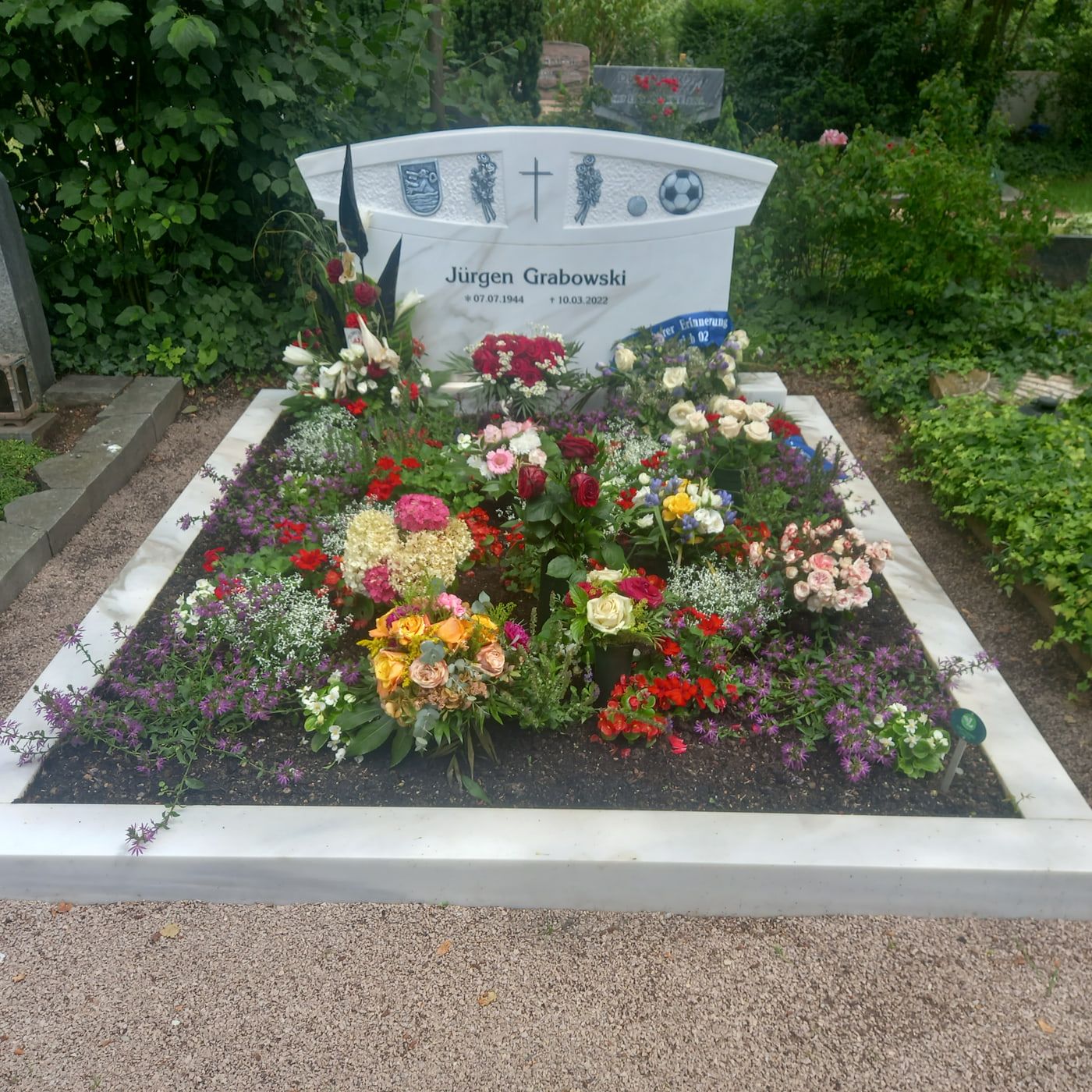 Weißes Grabmal Jürgen Grabowskis mit Kreuz, Wappenzeichen und Fußballmotiven vor einem dicht bepflanzten Beet auf Friedhof Biebrich